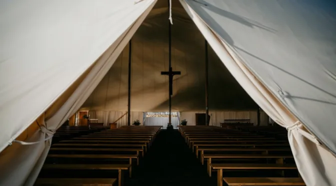 Interior view of an empty canvas revival tent with wooden benches aligned toward a central cross, symbolizing a sacred space prepared for worship, prayer, and the outpouring of God’s presence.